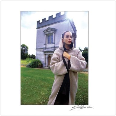 A woman in a long coat stands in front of a castle-like building.