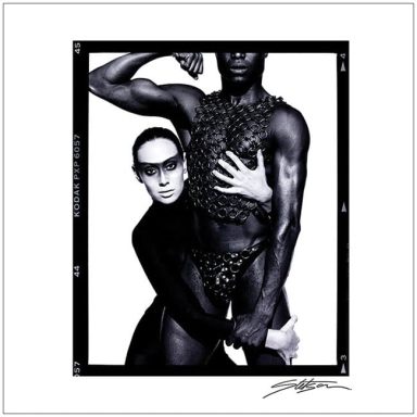 Two figures pose together; one in a black outfit and glasses, the other in a textured harness.