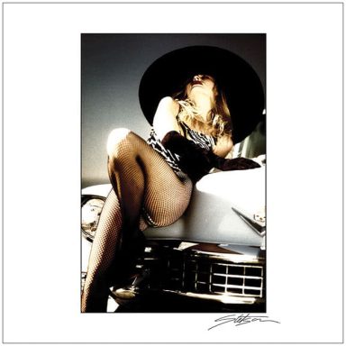 A glamorous woman in a black hat poses on a classic car, wearing a stylish outfit.