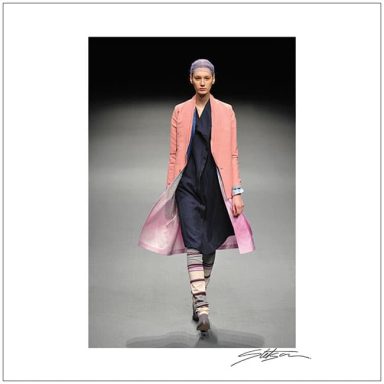 Model walking down the runway wearing a pink coat over a dark dress.