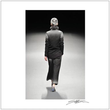 A model in a dark outfit walks away on a dimly lit runway.