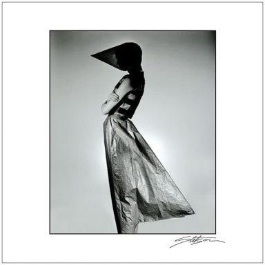 A person in a dramatic, conical hat and flowing dress posed against a minimalist background.