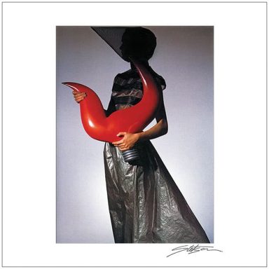 A figure holding a large, red, curving object, dressed in a dark garment against a neutral backdrop.