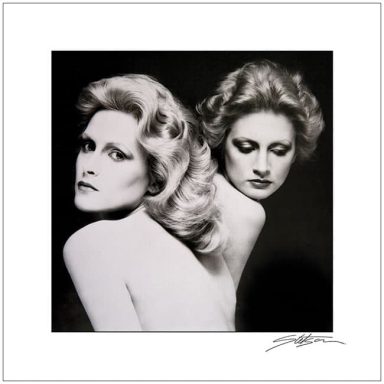 Black and white portrait of two women with stylized hair and makeup posed back-to-back.