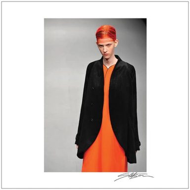 Model with short red hair wearing an orange dress and a black jacket on a runway.